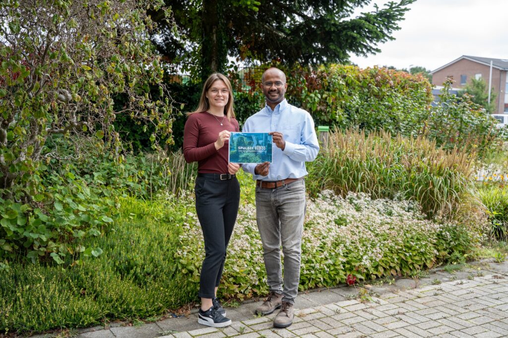 Two Spaleck employees are standing in front of a flower bed, holding the cover page of the new sustainability report